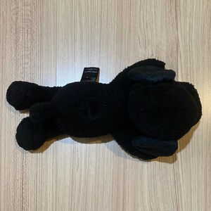 May include: A black plush toy in the shape of a dog, lying on its side. The toy has floppy ears and a small tail. The plush is made of a soft, textured material.