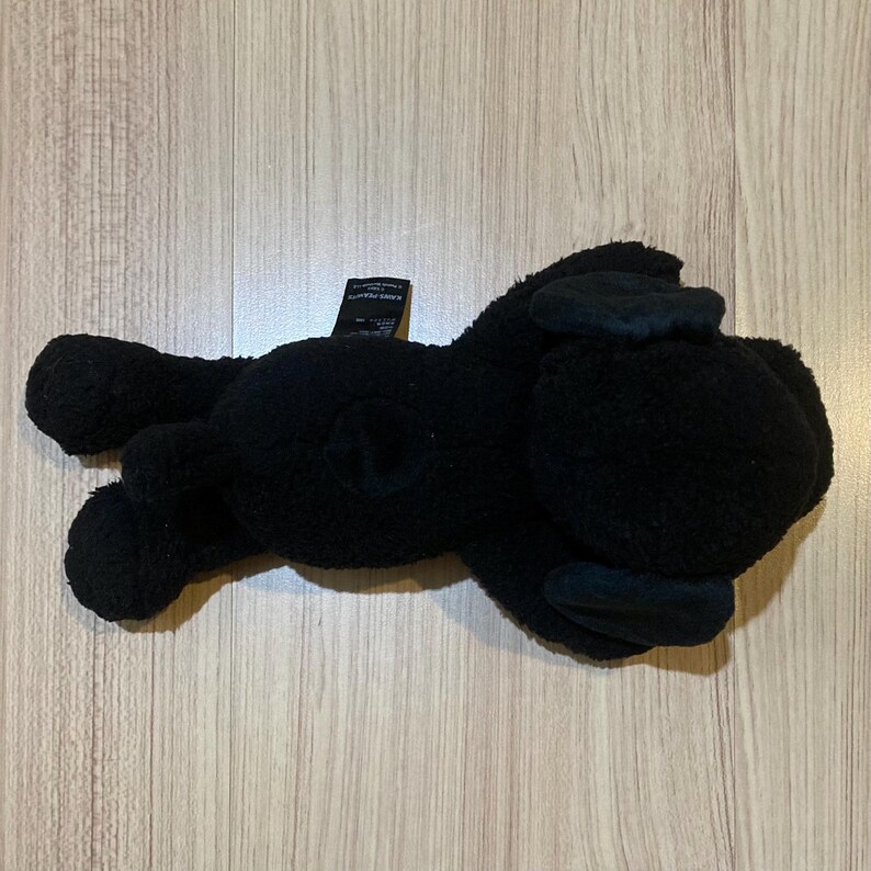 May include: A black plush toy in the shape of a dog, lying on its side. The toy has floppy ears and a small tail. The plush is made of a soft, textured material.