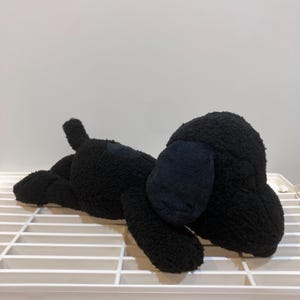 May include: A plush black dog toy with floppy ears and a small tail. The dog is lying down on a white surface, with its body made of a soft, textured material. The toy is designed to resemble a cartoon dog.