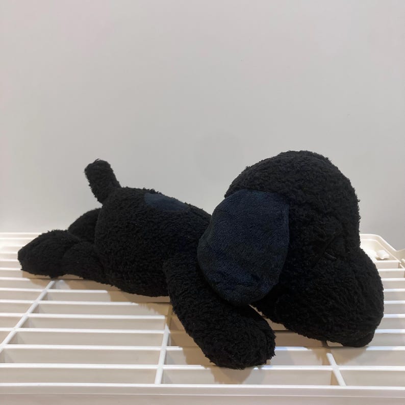 May include: A plush black dog toy with floppy ears and a small tail. The dog is lying down on a white surface, with its body made of a soft, textured material. The toy is designed to resemble a cartoon dog.