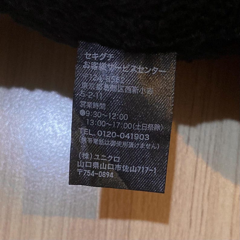 May include: A black tag with Japanese text, including company information and contact details. The tag also lists business hours and a phone number. The background is a dark, textured fabric.