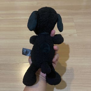 May include: A black plush Snoopy dog toy with floppy ears and a small green heart-shaped patch on its rear. The toy is held up by a person. The Snoopy toy has a tag attached to it.