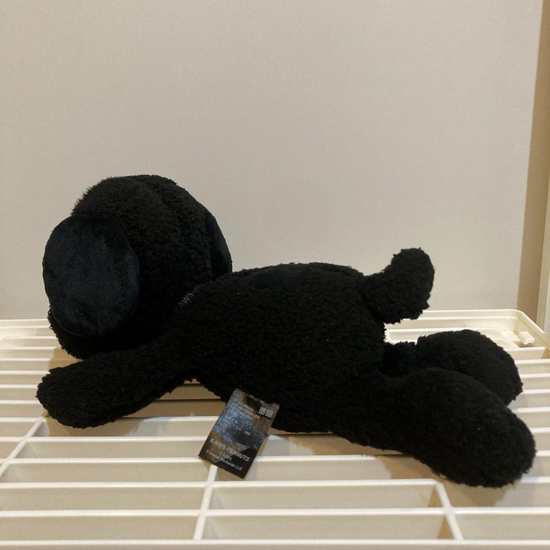 May include: A black plush dog toy lying on its side. The toy has a soft, textured appearance and a tag attached to its side. The dog's ears are floppy, and its tail is curled. The toy is resting on a white, slatted surface.