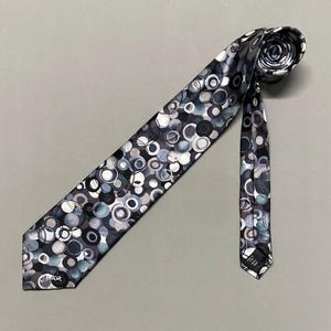 Może przedstawiać: Wzorzysty krawat z powtarzającym się wzorem nakładających się na siebie kół w odcieniach szarości, błękitu i bieli. Krawat jest zwinięty na górze i ma podpis na dole. Tło jest jednolicie szare.