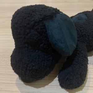 May include: A black plush dog toy with a textured, fuzzy surface. The dog has large, dark blue ears and a matching patch on its back. The toy is lying on a light brown wooden surface.