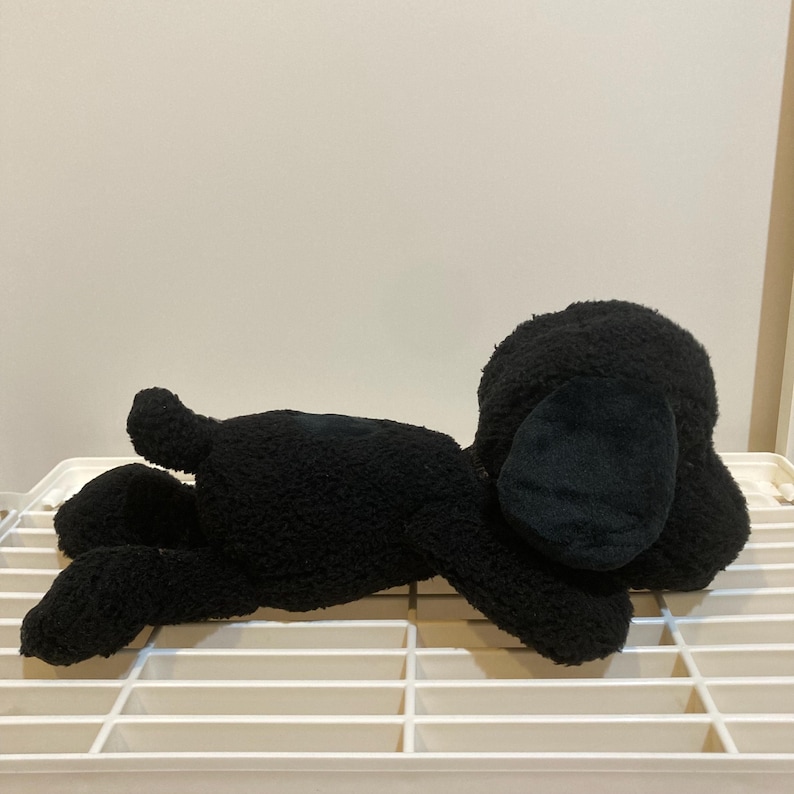 May include: A black plush dog toy in a lying down position. The toy has a textured, fuzzy exterior, with large floppy ears and a small tail. The dog is lying on a white, grid-like surface.
