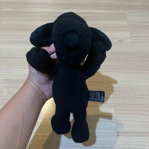 May include: A black plush toy in the shape of a creature with large ears and outstretched arms. The toy is made of a soft, textured material and has a tag attached to its side. The toy is being held up by a person.