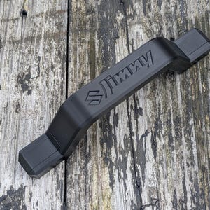 May include: A black car accessory, rectangular in shape, featuring the word "JIMMY" and a logo. The accessory is made of a solid material. It is designed for a vehicle and is shown on a weathered wooden surface. The item is likely a handle or trim piece.