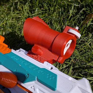 May include: A red sight attachment for a toy gun, with white and orange accents. The sight is mounted on a white, teal, and orange toy gun, resting on a bed of green grass. The sight has the text "A.S.O.N."