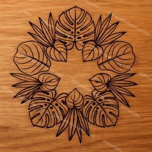 May include: A circular design of black tropical leaves, including monstera and palm fronds, is centered on a light brown wooden surface. The leaves are arranged in a symmetrical pattern, creating a decorative botanical motif.