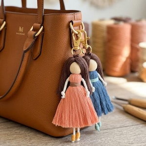 May include: Two doll-shaped bag charms made of macrame cord and wooden beads attached to a brown leather handbag. One doll wears a peach-colored dress with brown hair, and the other wears a blue dress with brown hair. Both have gold metal clasps.