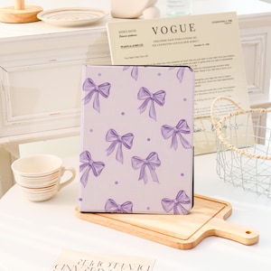 May include: A lavender tablet case with a pattern of purple bows and dots. The rectangular case is made of a smooth material. The design is feminine and decorative, suitable for protecting a tablet. The case is approximately 20 cm x 15 cm.