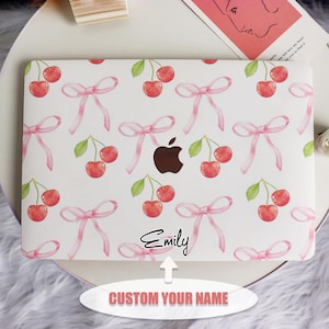 Può includere: Pelle per laptop bianca con un motivo di fiocchi rosa e ciliegie rosse. La pelle ha uno spazio per un nome personalizzato, che è mostrato come "Emily" nell'immagine.