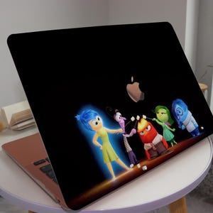 May include: A black laptop with a colorful sticker of the characters from the movie Inside Out. The sticker features Joy, Fear, Anger, Disgust, and Sadness. The characters are standing in a line and appear to be having a conversation. The laptop is on a white table.