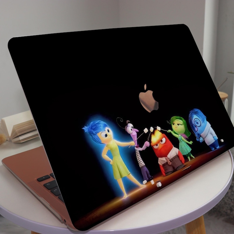 May include: A black laptop with a colorful sticker of the characters from the movie Inside Out. The sticker features Joy, Fear, Anger, Disgust, and Sadness. The characters are standing in a line and appear to be having a conversation. The laptop is on a white table.