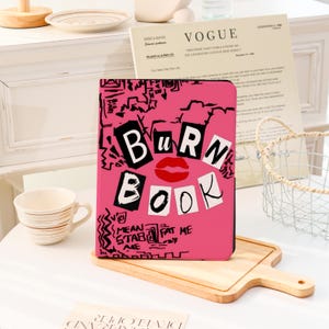 May include: A pink notebook with the words "BURN BOOK" in bold black letters, a red lip graphic, and additional text. The notebook is on a wooden cutting board.