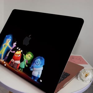 May include: A black laptop with a sticker of the characters from the movie Inside Out. The characters are Joy, Sadness, Anger, Disgust, and Fear. They are all smiling and looking at the camera. The laptop is closed and sitting on a white table.