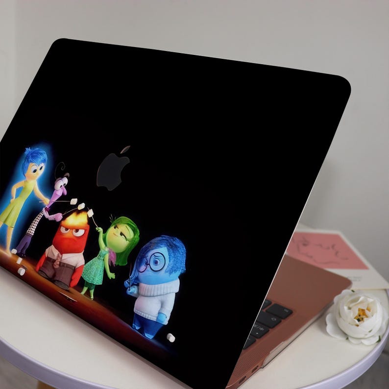 May include: A black laptop with a sticker of the characters from the movie Inside Out. The characters are Joy, Sadness, Anger, Disgust, and Fear. They are all smiling and looking at the camera. The laptop is closed and sitting on a white table.