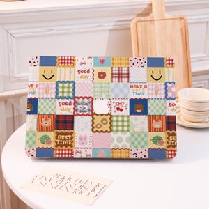 May include: A rectangular laptop case with a patchwork design. The case features various squares with images of a smiley face, ice cream, a bear, and the words "good day" and "best time". The case is on a white surface.