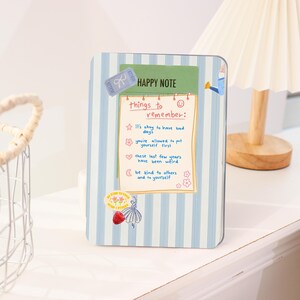 May include: A rectangular notebook with a blue and white striped pattern. The notebook has a green label that says "HAPPY NOTE" and a list of "things to remember". Decorative stickers include a bow, a watering can, and a ballerina.