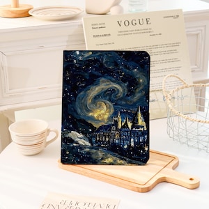 Könnte beinhalten: Ein dunkelblaues und goldenes Notizbuch mit einem Sternennacht- und Schlossdesign. Das Notizbuch wird auf einem Holzschneidebrett präsentiert. Eine weiße Tasse und eine Vogue-Zeitschrift befinden sich im Hintergrund.