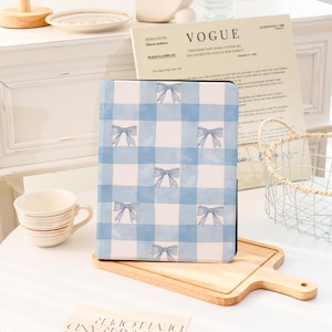 Op de afbeelding: Een notitieboek met een blauw en wit geruit patroon en strik illustraties. Het notitieboek ligt op een houten plank. Een witte mok en een exemplaar van het tijdschrift Vogue staan op de achtergrond.