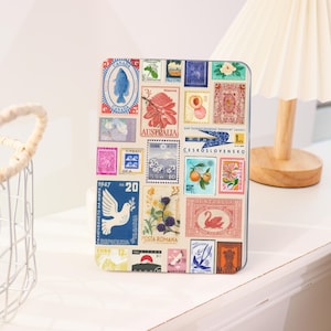 Puede incluir: Una caja metálica rectangular cubierta con un collage de coloridos sellos postales. Los sellos presentan varios diseños, incluyendo flores, pájaros y frutas, con textos como "Australia" y "Canada". La caja está sobre una superficie blanca.