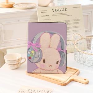 Peut inclure: Une housse de tablette avec un lapin de dessin animé portant un casque et des nœuds roses. Le lapin a un visage et des oreilles rose clair, avec un casque gris décoré d'étoiles et d'un motif Hello Kitty. Le fond est violet clair.