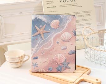Può includere: Una custodia per tablet con una scena di spiaggia, con una stella marina, conchiglie e onde nei toni del rosa, blu e beige. La custodia è su un tagliere di legno.
