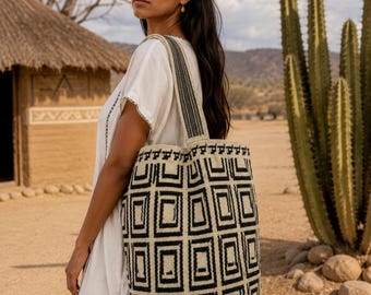 Puede incluir: Un bolso tote grande con un patrón geométrico repetitivo de cuadrados en negro y crema. El bolso tiene una correa de hombro tejida ancha a juego con un diseño lineal en negro y crema. La estructura es cilíndrica con base plana.