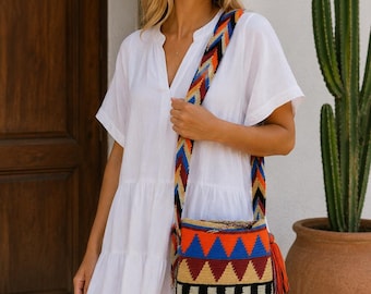 May include: A white short-sleeved tiered dress with a V-neckline paired with a cylindrical woven shoulder bag. The bag features a vibrant geometric pattern in orange, blue, black, and beige, with a matching wide strap and a decorative orange tassel.