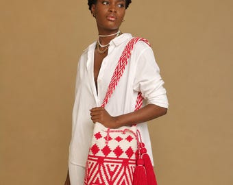 May include: A white button-down shirt and white shorts paired with a cylindrical bucket bag featuring a red and white geometric pattern. The bag has a long woven shoulder strap and two large red tassels hanging from the side with wooden beads.