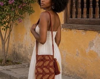 May include: A cylindrical woven bucket bag featuring a deep rust-red base with a repeating geometric chevron pattern in mustard yellow. The bag includes a long shoulder strap and a decorative dual-tone tassel in rust-red and mustard yellow.