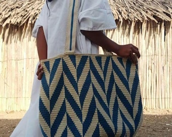 Puede incluir: Un bolso tote grande tejido con un patrón de chevron azul y beige. El bolso tiene forma redondeada y una correa ancha con rayas azules y beige. La persona lleva un vestido blanco.