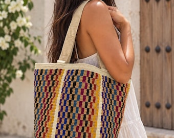 Puede incluir: Un bolso tote grande con un patrón tejido de rayas verticales en negro, rojo, azul y amarillo. El bolso tiene dos correas de hombro anchas de tela beige claro y un diseño estructurado de apertura superior con borde azul.