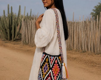 Puede incluir: Un bolso tipo cubo negro con un vibrante patrón geométrico de diamantes multicolor en tonos rosa, amarillo, morado y azul. El bolso incluye una correa de hombro larga, trenzada y multicolor, además de una gran borla de hilo multicolor.