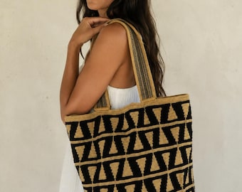 Puede incluir: Un bolso tote grande con un patrón geométrico repetitivo de triángulos negros sobre un fondo beige. El bolso está construido con una textura tejida e incluye dos correas de hombro a juego con rayas negras y beige para llevar el artículo.
