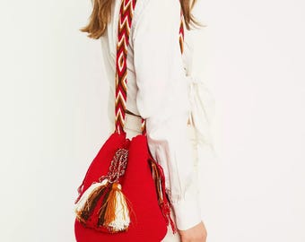 May include: A red woven shoulder bag with a decorative strap featuring a geometric pattern in red, white, and gold. The bag has a tassel and is worn over the shoulder.