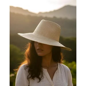 Puede incluir: Un sombrero fedora de paja en color beige claro con una copa estructurada y un ala ancha ligeramente inclinada hacia abajo. El sombrero presenta una textura tejida en toda su superficie, lo que le confiere un aspecto natural. Incluye una copa pellizcada.