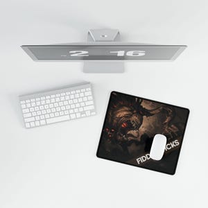May include: A computer setup featuring a silver monitor displaying the numbers "2 16", a white keyboard, and a black mousepad. The mousepad has a graphic of a monster with red eyes and the text "FIDDCKS". A white mouse rests on the mousepad.