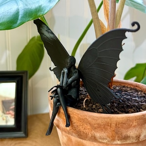 Könnte beinhalten: Eine schwarze Feenfigur mit Flügeln sitzt auf dem Rand eines Terrakotta-Blumentopfs. Die Fee hält ihre Hände vor ihrem Gesicht zusammen. Der Topf ist mit Erde gefüllt und eine grüne Pflanze wächst darin.