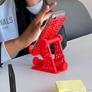 May include: A red 3D printed phone stand with a geometric design. The stand is holding a smartphone in a vertical position.