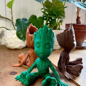 Może przedstawiać: Trzy doniczki Groot wydrukowane w 3D w różnych kolorach: brązowym, zielonym i brązowym. Zielony Groot siedzi ze skrzyżowanymi nogami, podczas gdy dwa brązowe Grooty siedzą z wyciągniętymi nogami.
