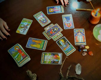 May include: A tarot card reading spread laid out on a wooden table. The cards are colorful and feature illustrations of people and symbols. There are also a few small crystals and a lit candle on the table.