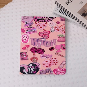 May include: Pink tablet case with a collage of graphics and text. The design includes hearts, dice, a ghost face, and the words "Bad Girls," "Bitch," and "Slut!" The case also features a book and a perfume bottle.