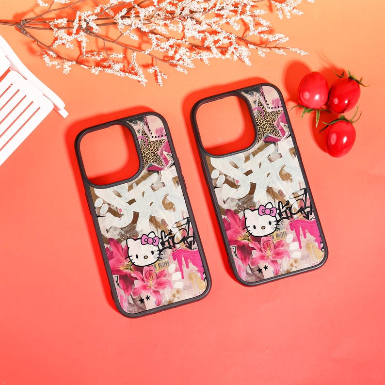 以下が含まれることがあります： 黒いフレームの2つの携帯電話ケース。Hello Kittyのグラフィック、ピンクの花、白いグラフィティ風のテキスト、ヒョウ柄の星がピンクの背景に描かれています。ケースはコーラル色の表面にあります。