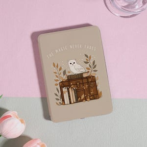 May include: A beige e-reader case featuring a white owl perched on a stack of books atop a brown vintage suitcase. The text THE MAGIC NEVER FADES is printed in white above the owl, surrounded by stylized brown and green leaves and fairy lights.