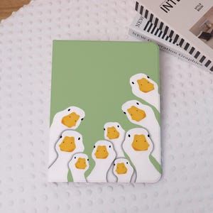 Op de afbeelding: Een groene tablethoes met een ontwerp van witte ganzen met gele snavels. De ganzen zijn rond de randen van de hoes geplaatst, met een lichtgroene achtergrond. Het ontwerp creëert een speelse rand.