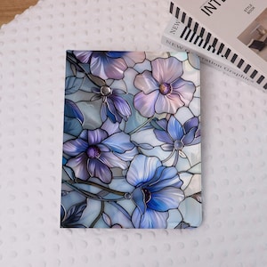 Peut inclure: Une couverture de carnet rectangulaire avec un motif floral en vitrail. Le motif présente de grandes fleurs stylisées dans des tons de bleu, de violet et de blanc, avec des détails complexes et une surface réfléchissante. La couverture est posée sur un fond blanc texturé.