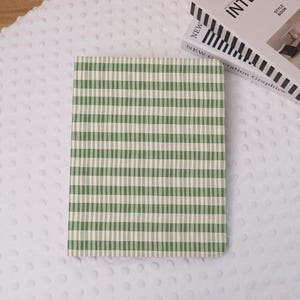 Op de afbeelding: Een notitieboek met een groen en wit gestreepte kaft ligt op een witte, getextureerde ondergrond. De kaft heeft een patroon van verticale strepen. Een boek met de titel "INTERIOR" is gedeeltelijk zichtbaar.